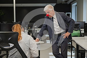 Elderly Caucasian man wearing a clown nose swears at his subordinate in the office.