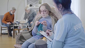 Elderly Caucasian woman talking with volunteer and knitting. Young girl supporting mature female retiree in nursing home
