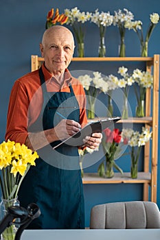 An elderly confident sincere smiling male florist holds folder for recording orders.
