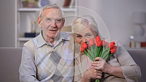 Elderly couple hugging at home, woman holding bunch of tulips, anniversary gift