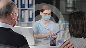 elderly couple in medical masks on face in consultation with female doctor during virus and infection discussing health