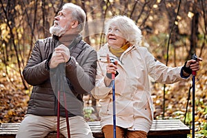 Elderly couple resting while sitting on bench with nordic walking poles, smiling