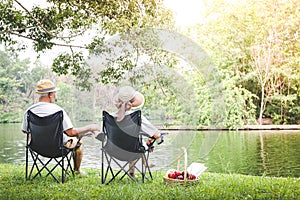 An elderly couple sit on a black chair in a shady garden and have a picnic basket for bread and fruit.