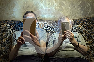 An elderly couple sit on the couch with gadgets, look at them close their faces.
