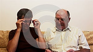 An elderly couple sits on a sofa at home and try on glasses for vision, consulting each other.