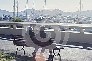 An elderly couple is sitting on a bench among white expensive yachts and mountains on a sunny day.