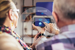 Elderly couple sitting comfortably on a sofa with their backs holiding remote control home system