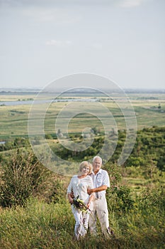 Elderly couple in the white linen dress standing outdoors, in the hands a bouquet of flowers, against the background of the field