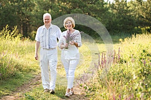 Elderly couple in the white linen dress walking outdoors, in the hands of a bunch of flowers, smiles on their faces, and on the ba