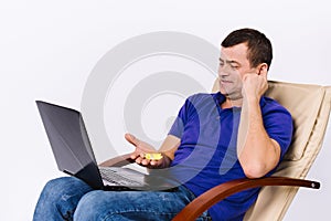 An elderly deaf man in a leather chair with a laptop and a smile shows a hearing aid at the camera. Online health