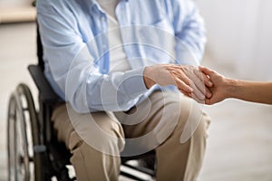 Elderly disabled people support. Young female holding hand of handicapped senior man in wheelchair indoors, closeup