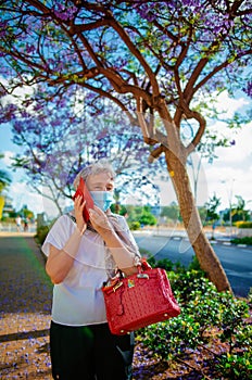 Elderly elegant lady in blue protective mask talking on a mobile phone under a purple jacaranda tree.