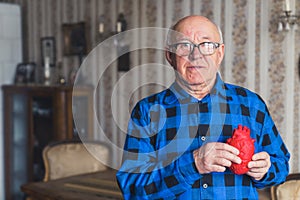 Elderly European bald senior man in blue flannel shirt holding artificial fake heart prop in front of his chest. Old