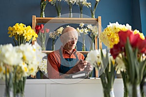 An elderly European man manager in flower shop arranges delivery using smartphone.