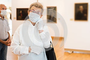 Elderly European woman  in mask protecting against covid examines paintings on display in hall of art museum