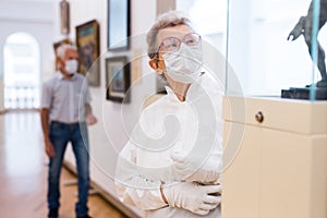 Elderly European woman  in mask protecting against covid examines paintings on display in hall of art museum