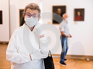 Elderly European woman  in mask protecting against covid examines paintings on display in hall of art museum