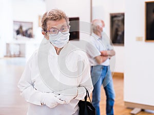 elderly European woman in mask protecting against covid examines paintings on display in hall of art museum