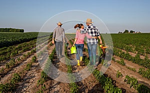 Elderly farmer and young couple walking in vegetable field with gardening equipment