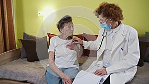 Elderly female doctor in white lab coat, medical mask makes medical examination of senior woman at home during