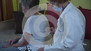 Elderly female doctor in white lab coat, medical mask makes medical examination of senior woman at home during