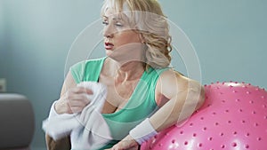 Elderly female leaning on fitness ball fanning herself with towel, exhaustion