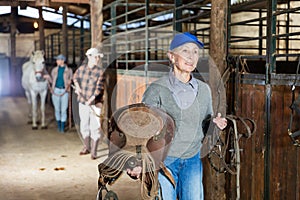 Elderly female rancher walking in stable with saddle in hands