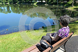 Older woman sits on bench with oxygen tank looking at pond photo