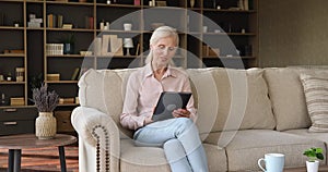 Elderly female sit on sofa spend leisure use digital tablet