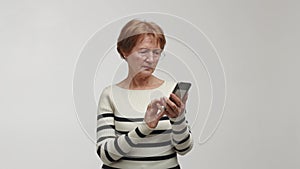 An elderly focused woman squints, has a poor eyesight and wears eyeglasses, holds a smartphone, brings screen closer to