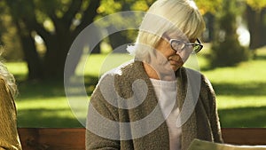 Elderly friends sitting apart after conflict, prying information in newspaper