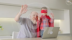 Elderly grandfather with gray beard, together with his grandson-a boy in red hero raincoat, sitting at table in living