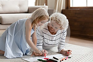 Elderly grandma teach little granddaughter drawing lying on warm floor