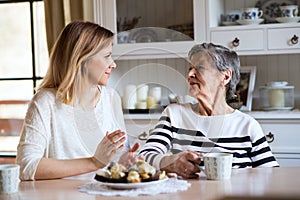 An elderly grandmother with an adult granddaughter sitting at the table at home, eating cakes.