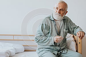 Elderly with gray beard and hair sits in pajamas on the bed with glasses in hand