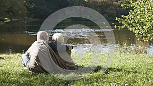 Elderly gray haired couple siting on the bench near the river on autumn day covered with blanket. Romance in old age