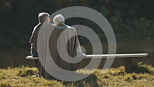 Elderly gray haired couple siting on the bench near the river covering with blanket on autumn day. Romance and old age