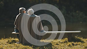 Elderly gray haired couple siting on the bench near the river covering with blanket on autumn day. Romance and old age