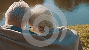 Elderly gray haired couple siting on the bench near the river covering with blanket on autumn day. Romance and old age