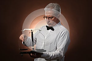 Elderly gray-haired man 50s, in white shirt, glasses and bow tie weighing something on scales with kettlebells