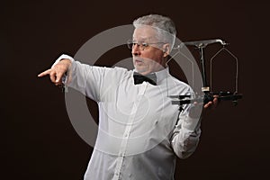 Elderly gray-haired man 50s, in white shirt, glasses and bow tie weighing something on scales with kettlebells