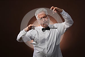 Elderly gray-haired man 50s, in white shirt, glasses and bow tie weighing something on scales with kettlebells