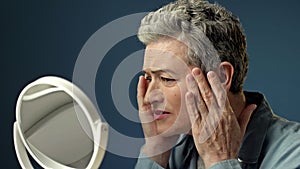 An elderly gray-haired woman carefully examines her reflection in the mirror, lightly touching the wrinkles on the skin