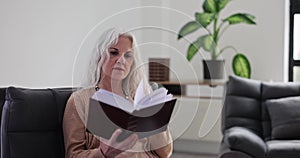 Elderly gray haired woman is relaxing on sofa and reading book