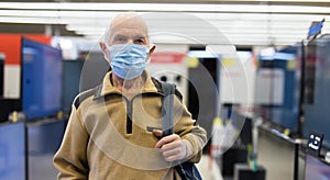 elderly grayhaired man pensioner in antiviral mask choosing modern digital televisors in showroom of digital goods store