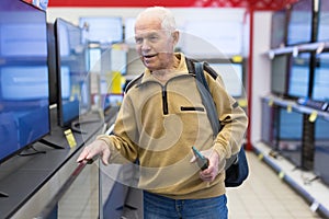 Elderly grayhaired man pensioner looking counter with modern digital televisors in showroom of digital goods store