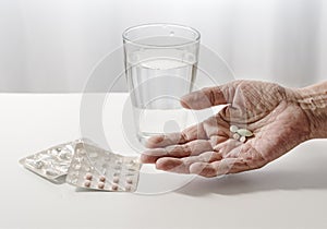 elderly hand of a senior is holding pills, water glass and blister pack on the white table, light background with copy space