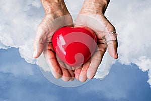 Elderly hands with wound holding red heart heart on blue sky and white clouds