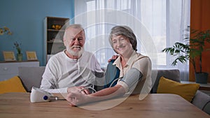 elderly happy couple looking at camera while taking blood pressure, husband using blood pressure monitor for wife with