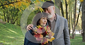 An elderly and happy couple are smiling and hugging in warm weather in a cozy autumn city park. Senior woman with leaves